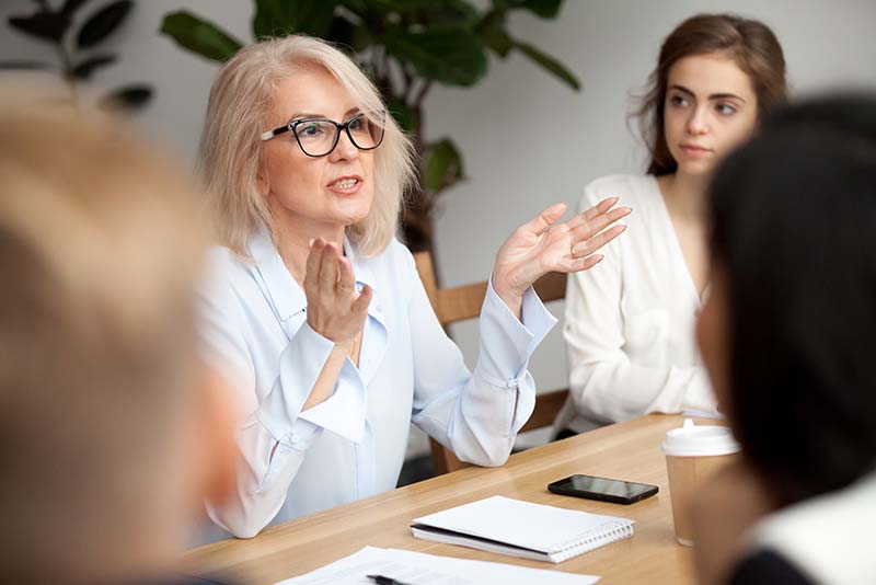 Older woman with white hair shares Foresee Now philosophy with her coworkers at leadership meeting